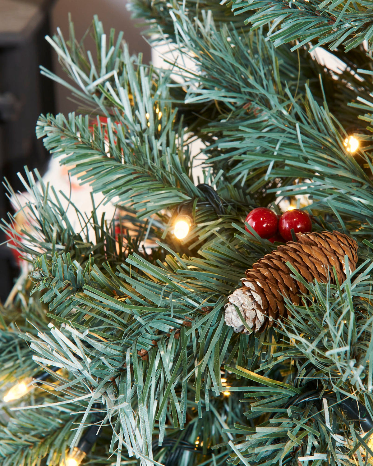 A pre-lit Scandinavian spruce Christmas tree with frosted green PVC branches, warm white LED lights, natural pinecones, and red berries. Positioned in a festive indoor setting with Christmas presents and a seasonal sign, capturing the rustic charm of a snow-dusted Nordic forest.