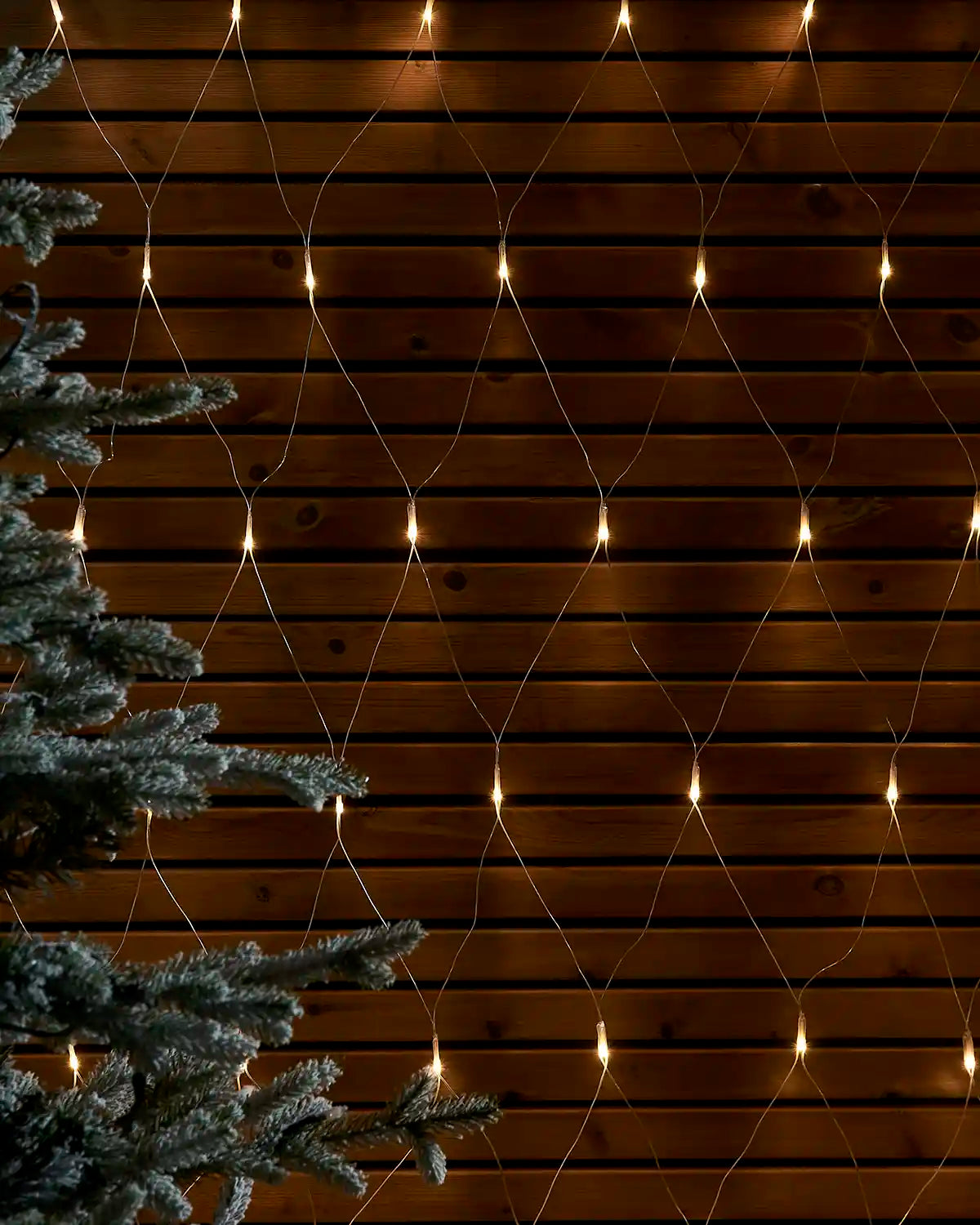 Warm white net lights displayed in a diamond pattern against a wooden wall beside a frosted tree.