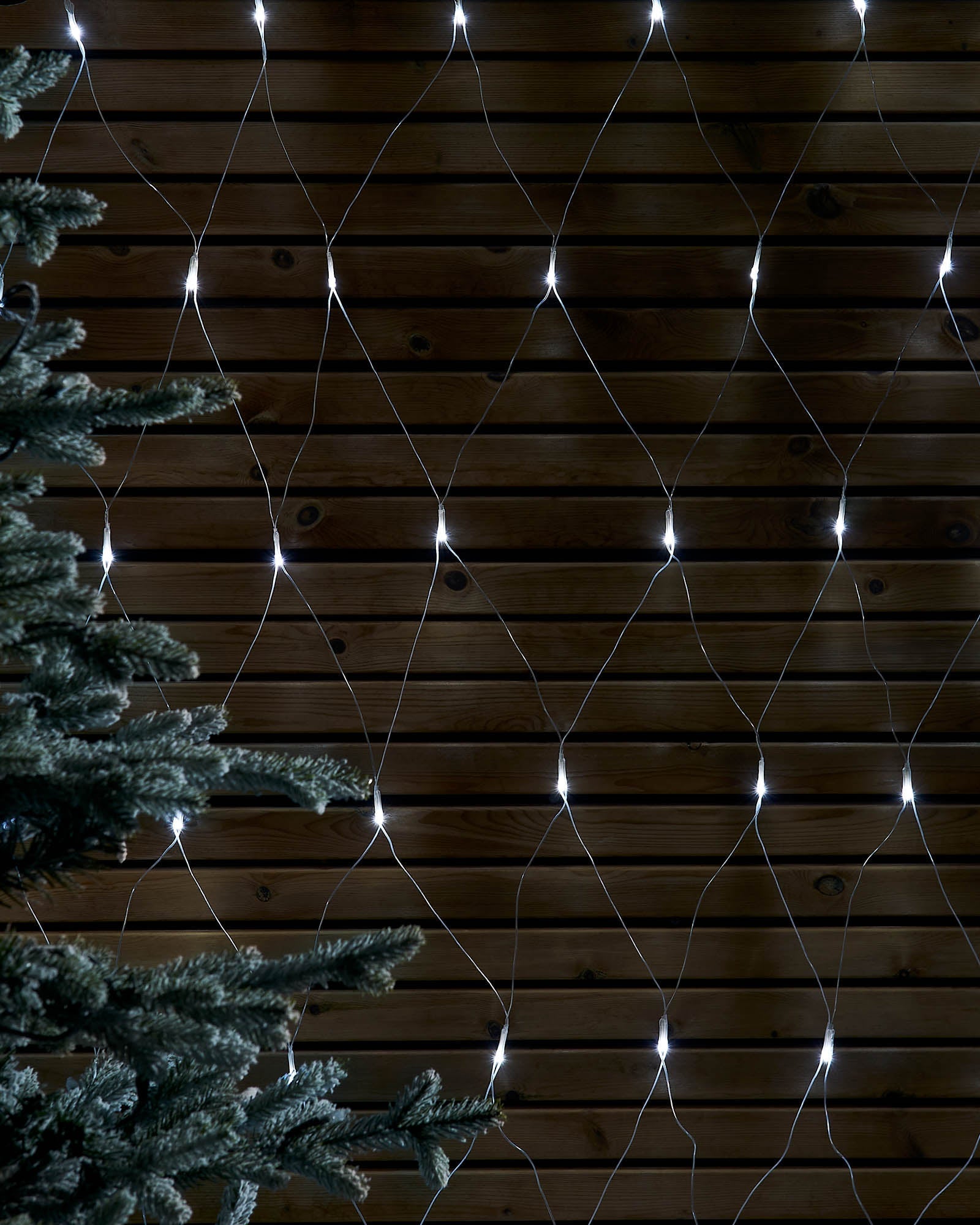 White net lights displayed in a diamond pattern against a wooden wall beside a frosted tree.