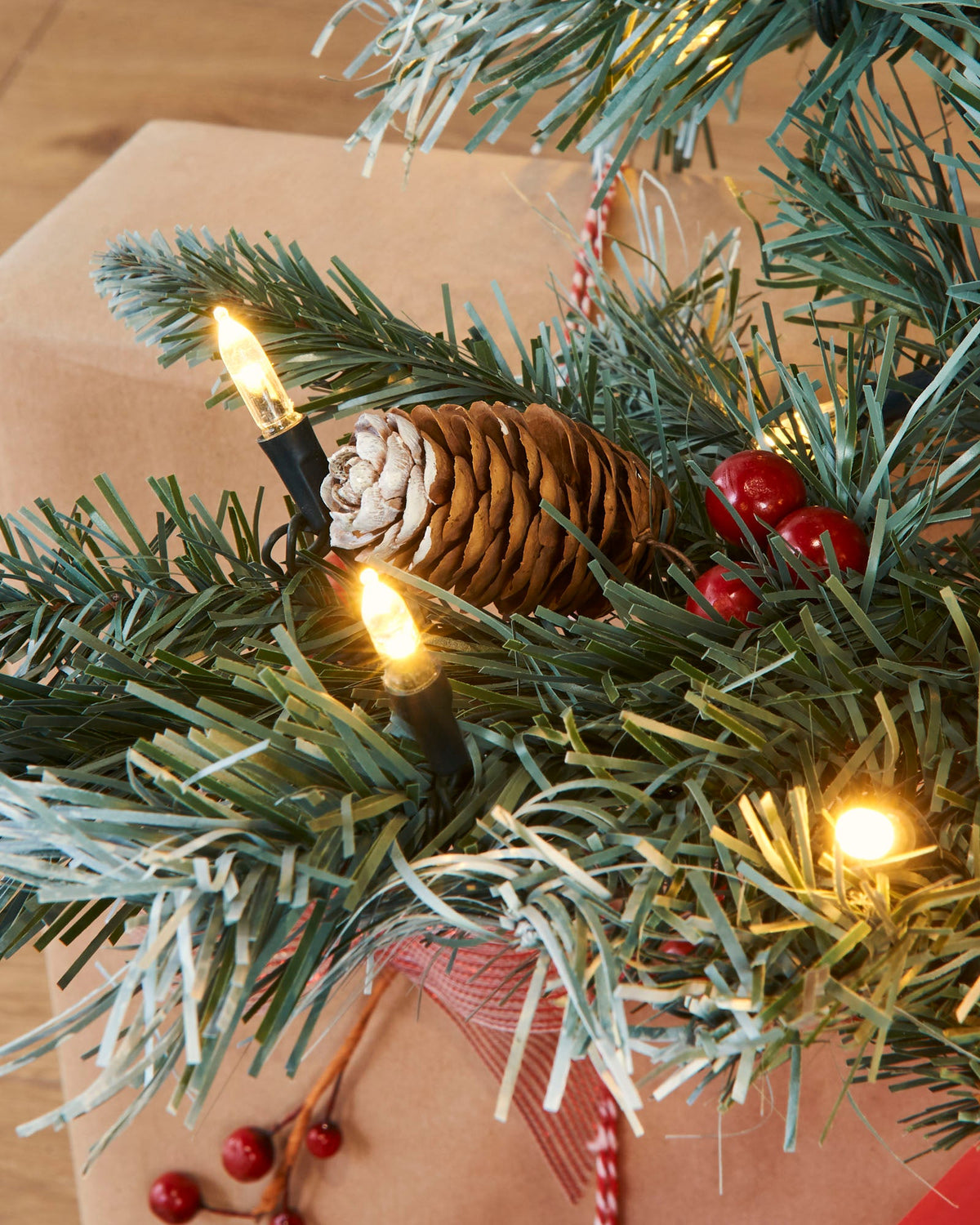 A pre-lit Scandinavian spruce Christmas tree with frosted green PVC branches, warm white LED lights, natural pinecones, and red berries. Positioned in a festive indoor setting with Christmas presents and a seasonal sign, capturing the rustic charm of a snow-dusted Nordic forest.