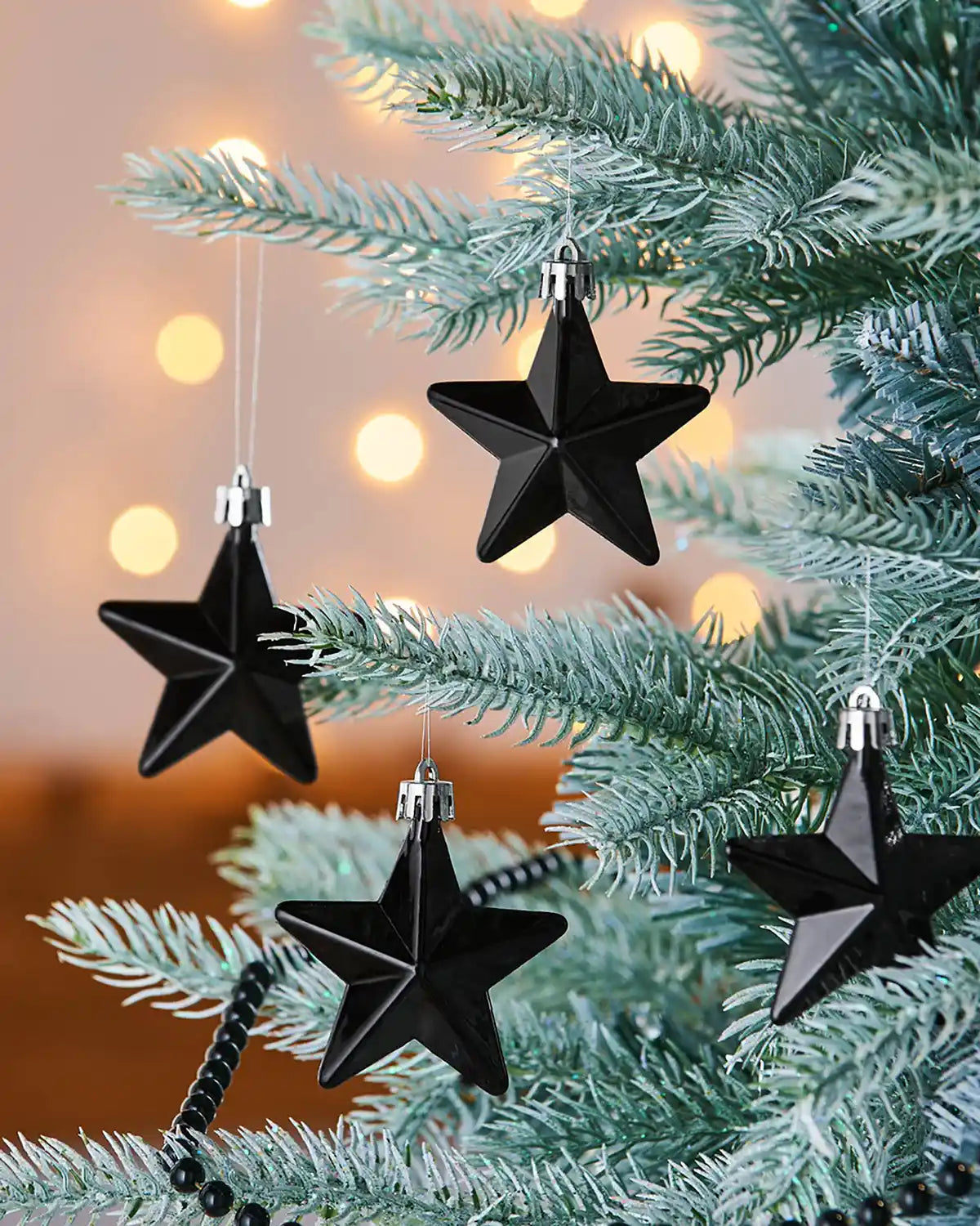 4 gloss Black star hanging baubles on a snow flocked Christmas tree with blurry lights in background 
