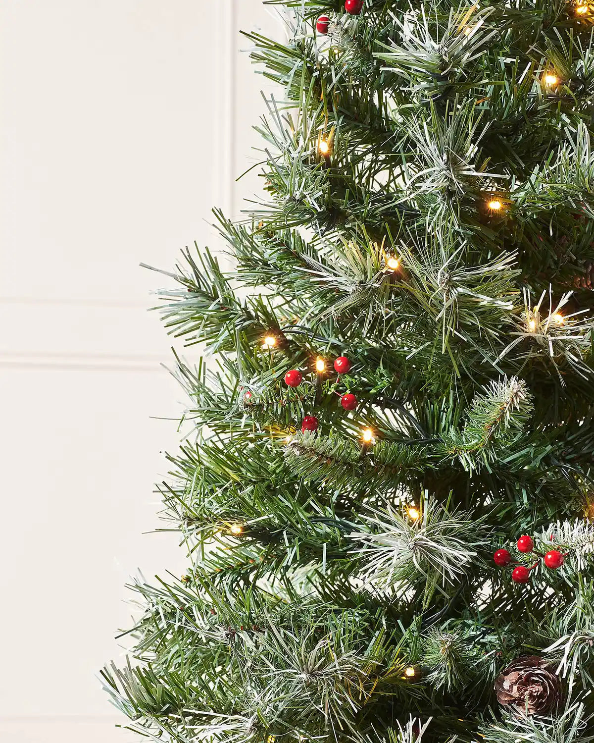 Close-up of frosted green branches with red berries, pinecones and warm white lights