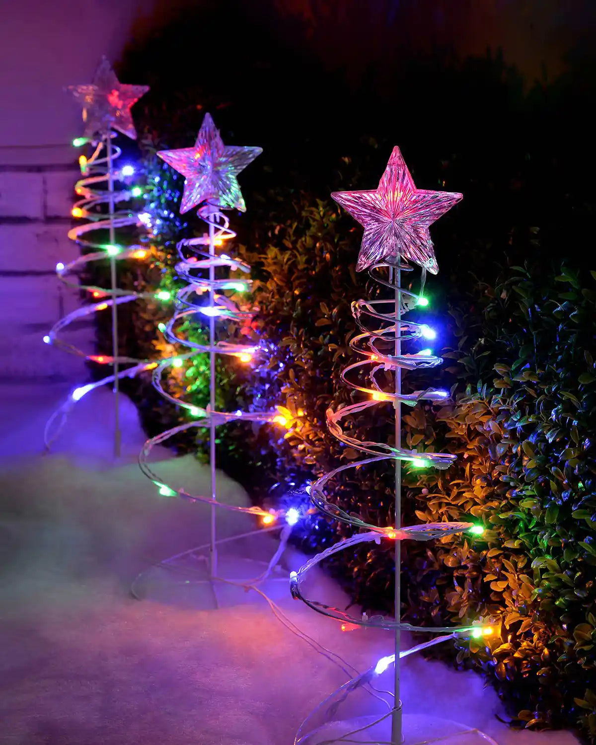 Multi-coloured spiral tree lights displayed outdoors beside a garden border