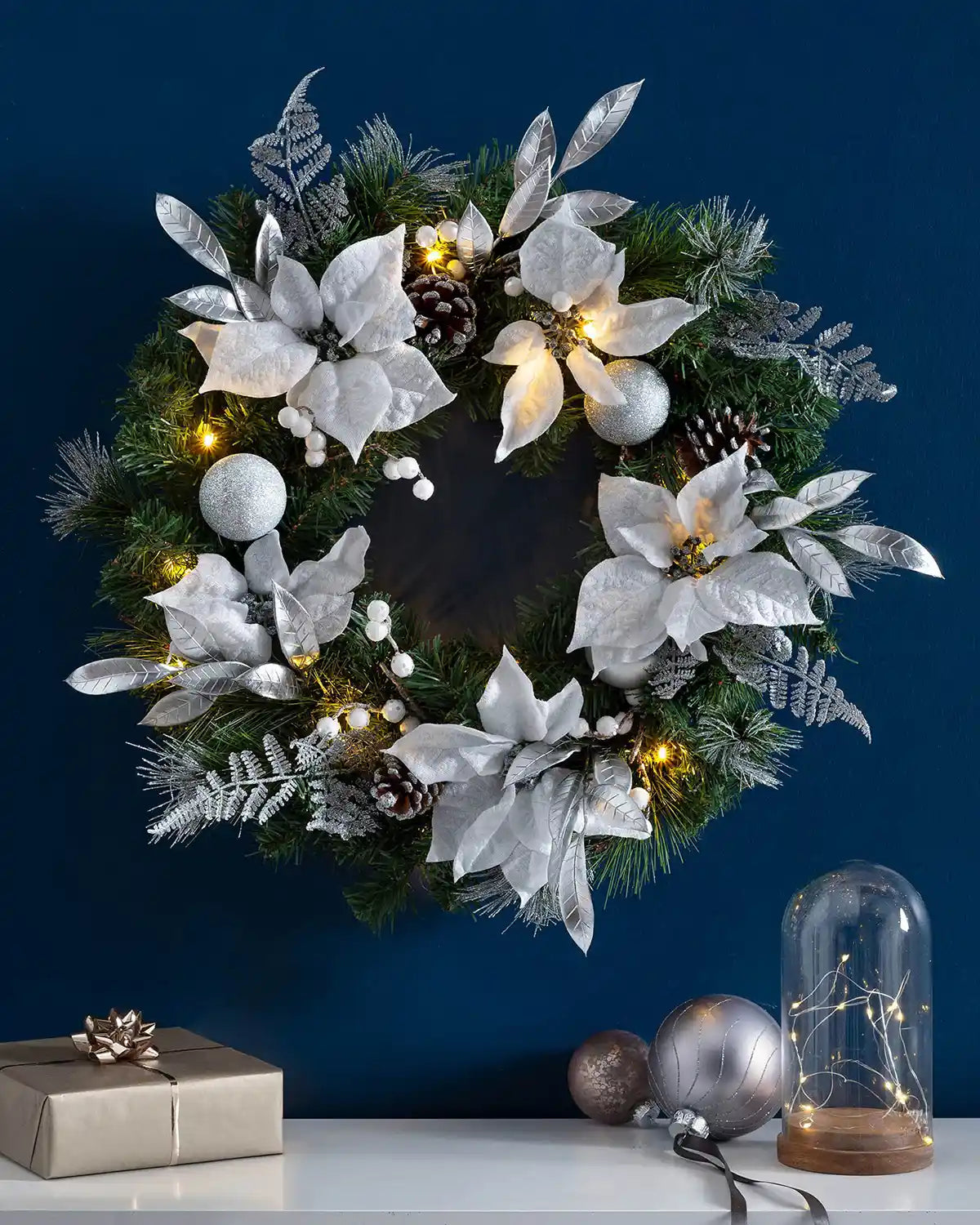 Silver and white pre-lit Christmas wreath styled on a blue wall with warm white lights
