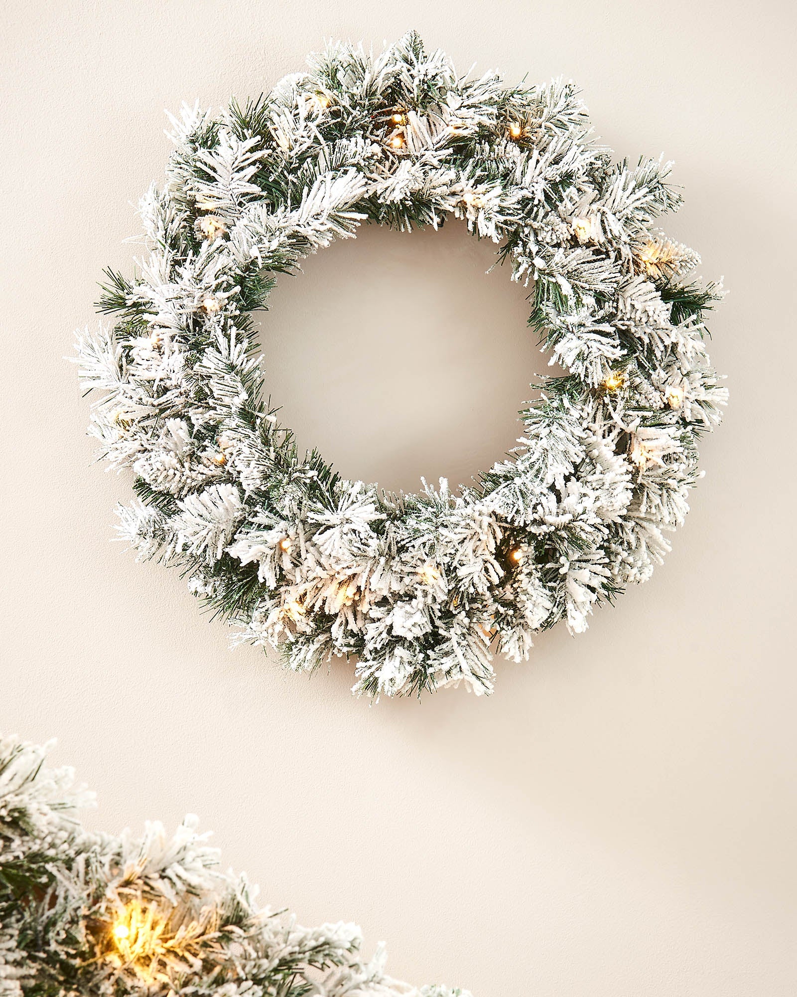60cm snow-flocked spruce wreath with warm white lights displayed on a plain wall, with matching garland partly visible below