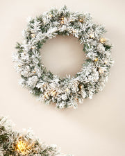 60cm snow-flocked spruce wreath with warm white lights displayed on a plain wall, with matching garland partly visible below