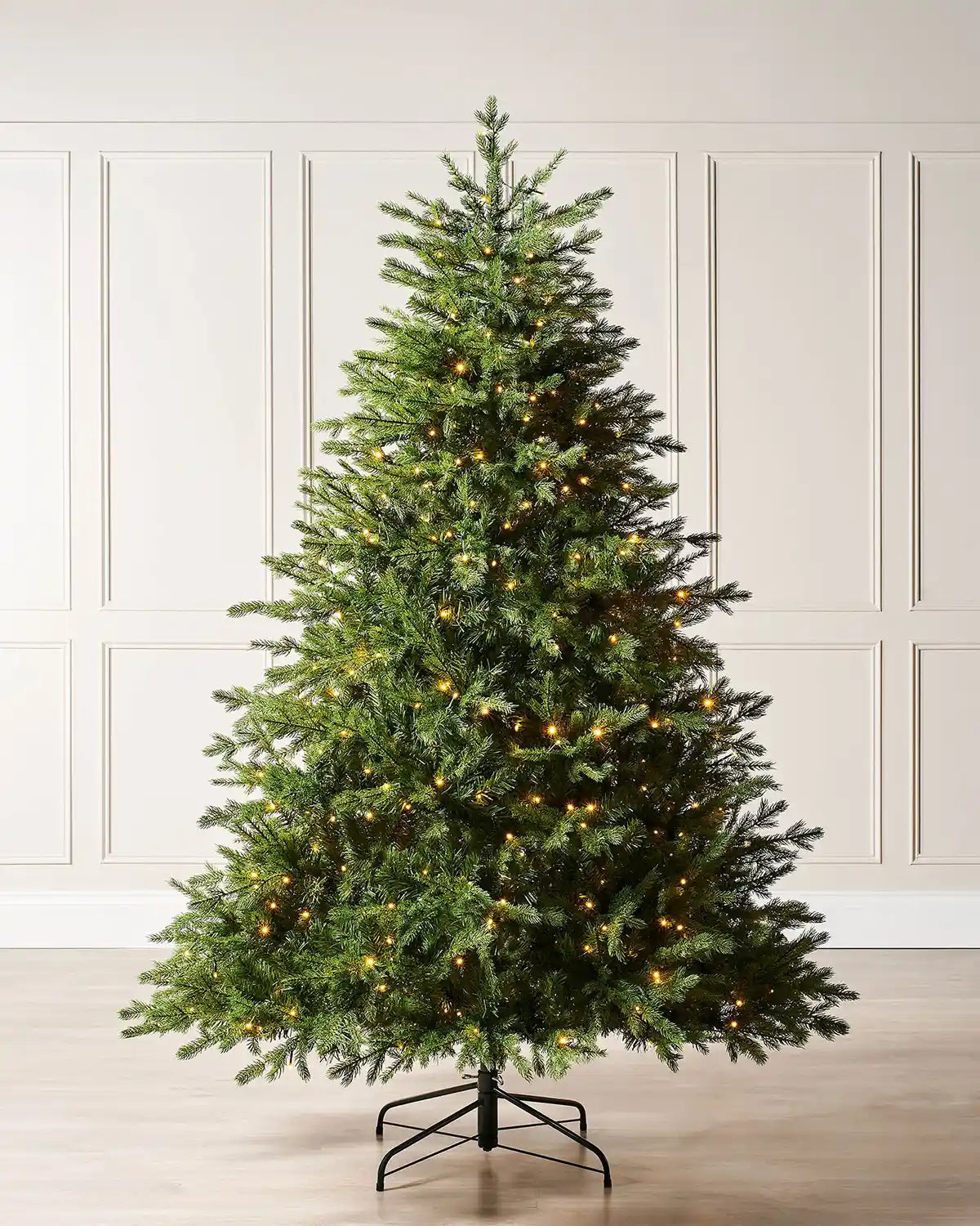 6ft pre-lit Windsor Fir Christmas tree displayed in a room setting with cream panelled wall, full green foliage, warm white lights and a foldable black metal stand