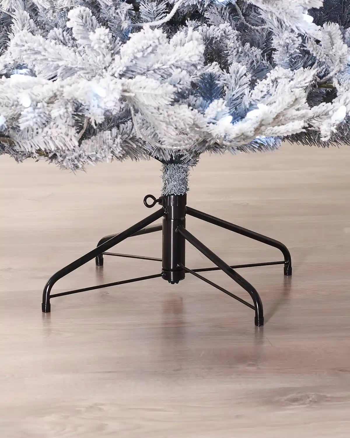 Base detail of 6ft snow flocked spruce Christmas tree showing snowy lower branches, cool white lights and metal tree stand