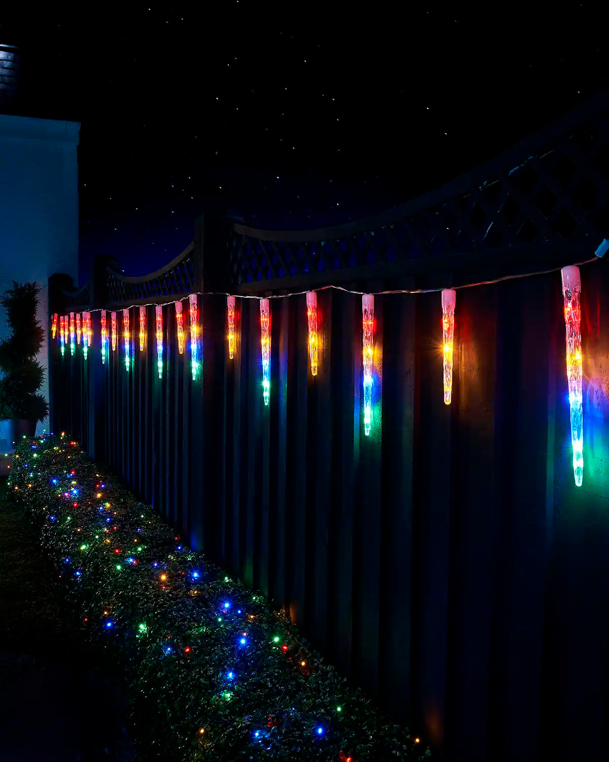 Multi-colour icicle drop lights hanging along a garden fence at night.