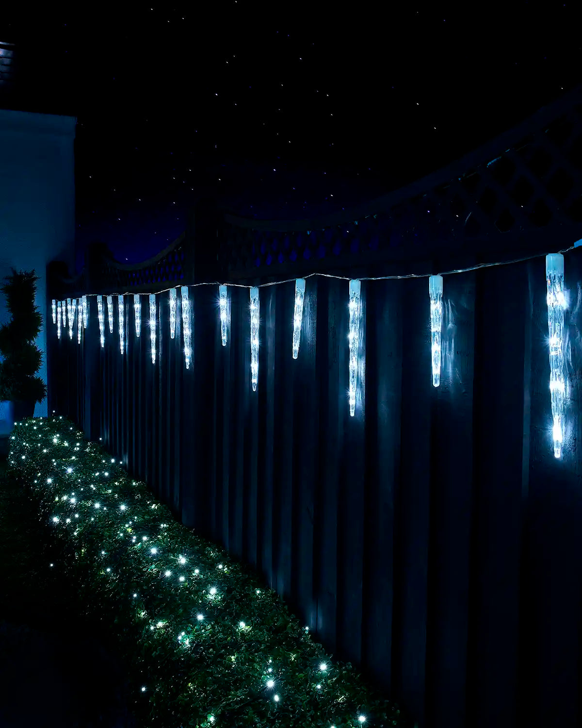 White icicle drop lights hanging along a garden fence at night.