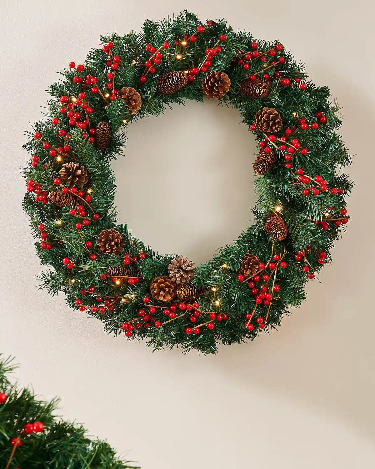 Extra thick pre-lit Christmas wreath styled on a wall with red berries, pinecones and warm white lights
