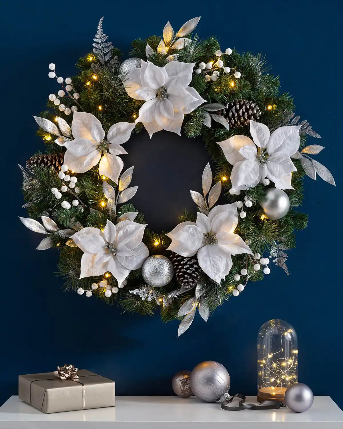 Extra thick silver and white pre-lit Christmas wreath styled on a blue wall with warm white lights