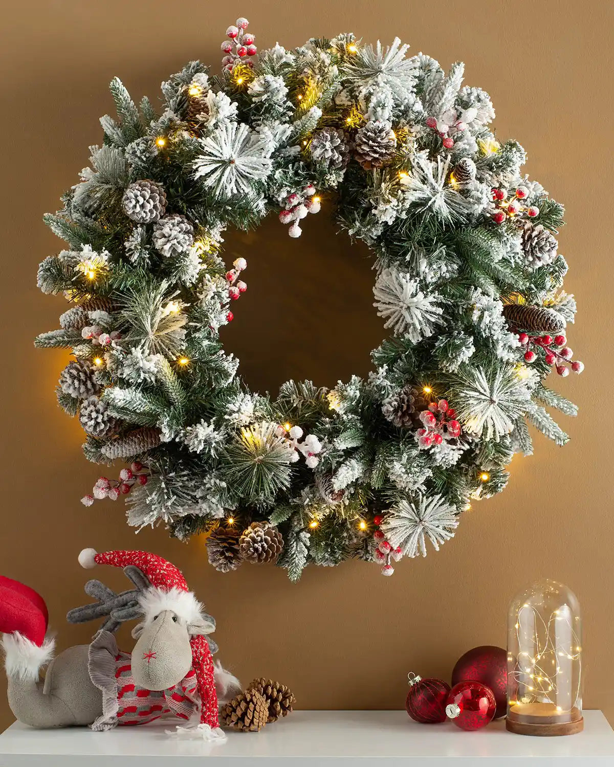 Snow-flocked pre-lit Christmas wreath styled on a wall with warm white lights, berries and pinecones
