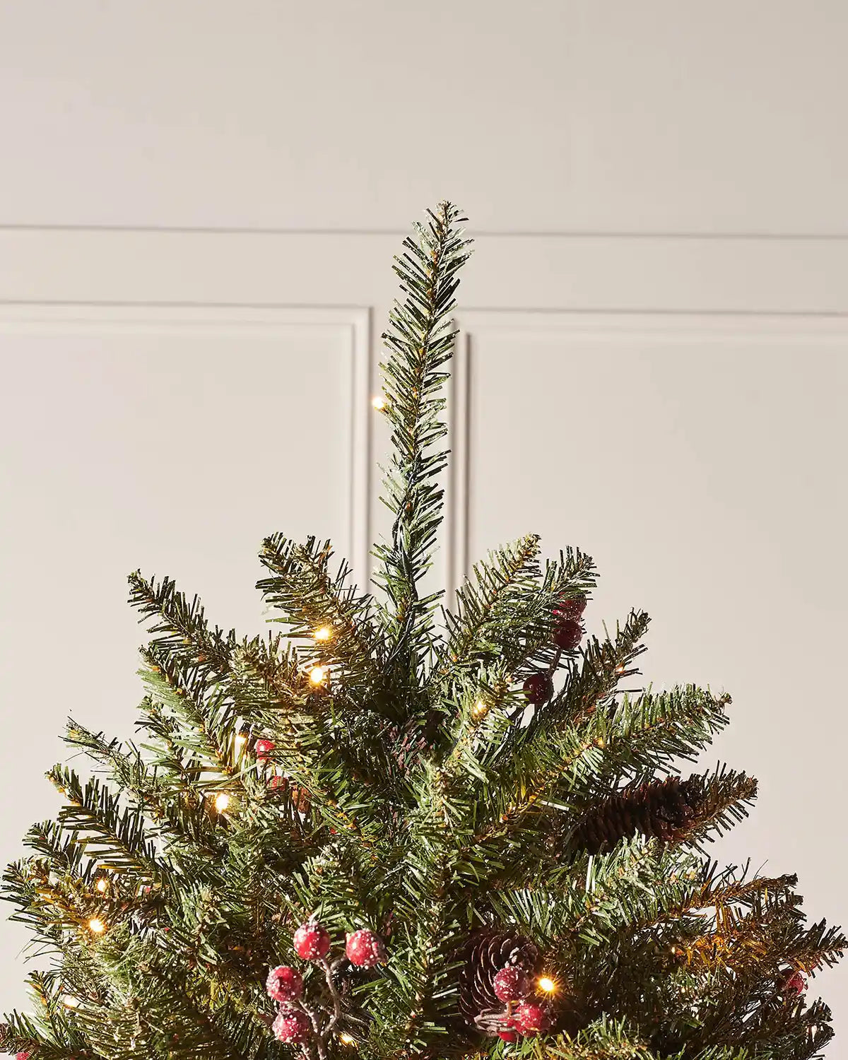 Top section of a frosted decorated Christmas tree with warm white lights, pinecones and red berries