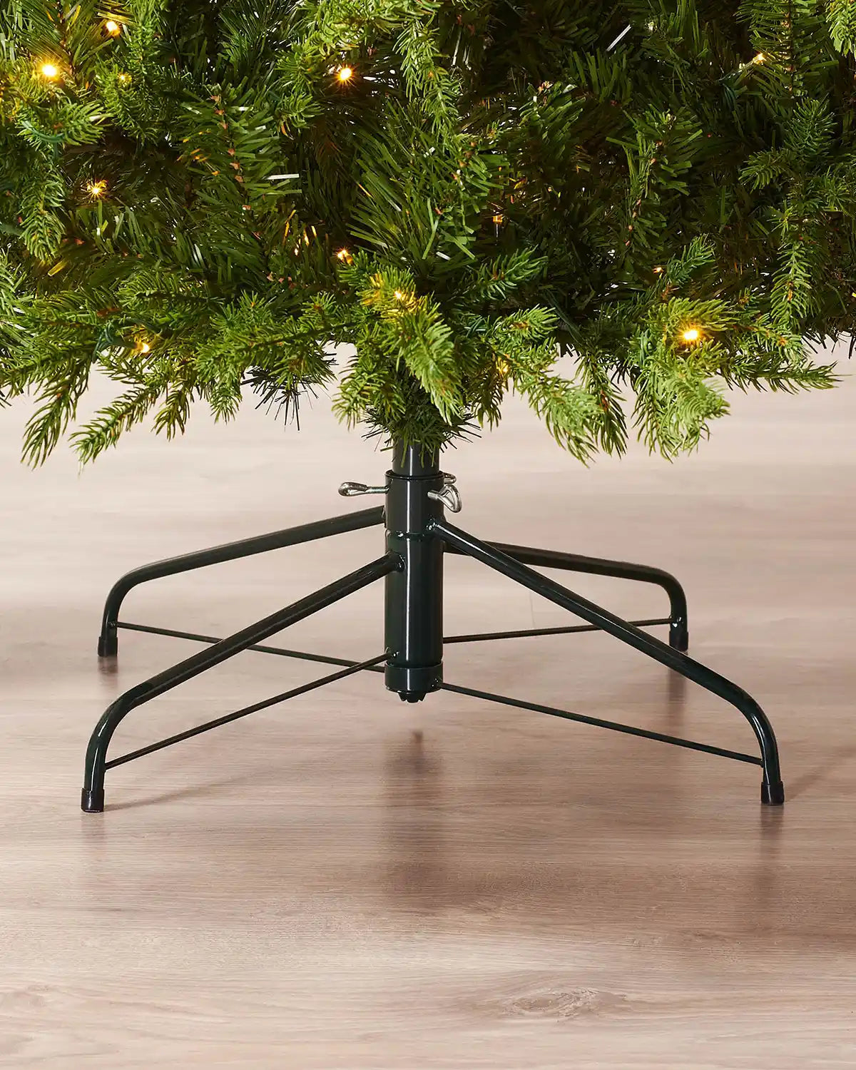 Base of 7ft pre-lit Windsor Fir Christmas tree showing full lower branches, warm white LEDs, central trunk and foldable black metal stand on a wooden floor