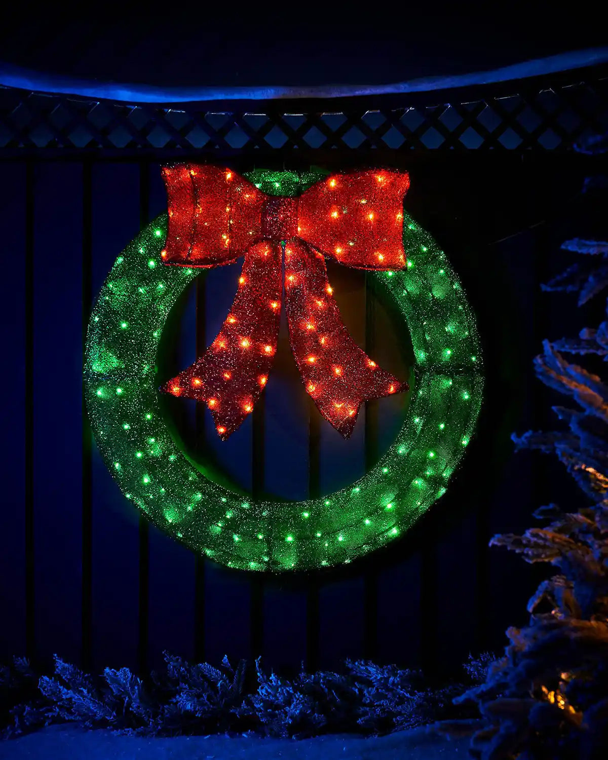 Pre-lit 85cm 3D wreath silhouette with green lights and a red bow on an outdoor fence.