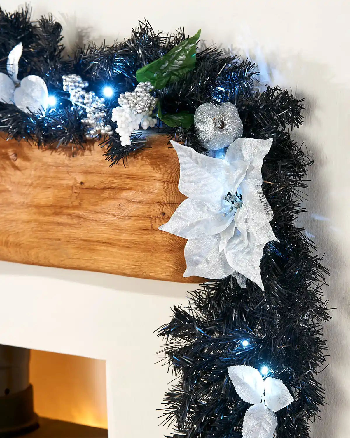 Black and silver decorated Christmas garland displayed on a mantelpiece with white poinsettia, silver bauble, berries and cool white lights