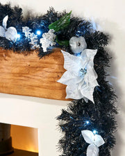 Black and silver decorated Christmas garland displayed on a mantelpiece with white poinsettia, silver bauble, berries and cool white lights