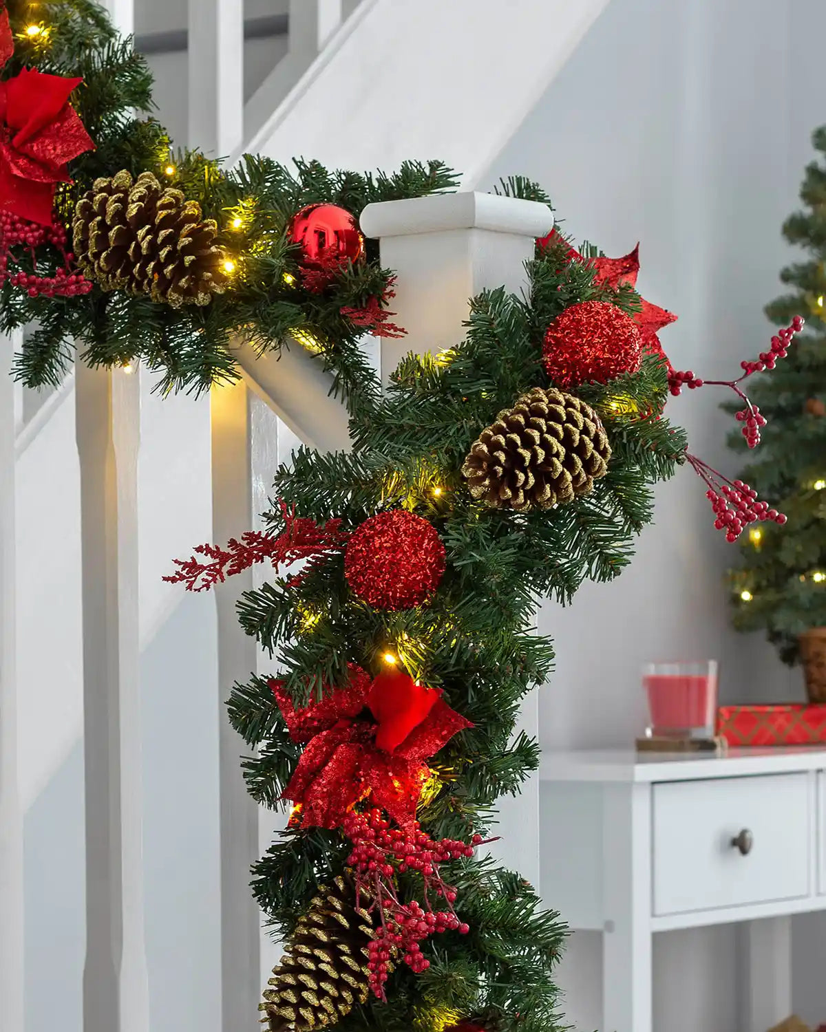 Pre-lit red decorated Christmas garland wrapped around a staircase banister with warm white LEDs, pinecones, baubles and berries