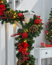 Pre-lit red decorated Christmas garland wrapped around a staircase banister with warm white LEDs, pinecones, baubles and berries