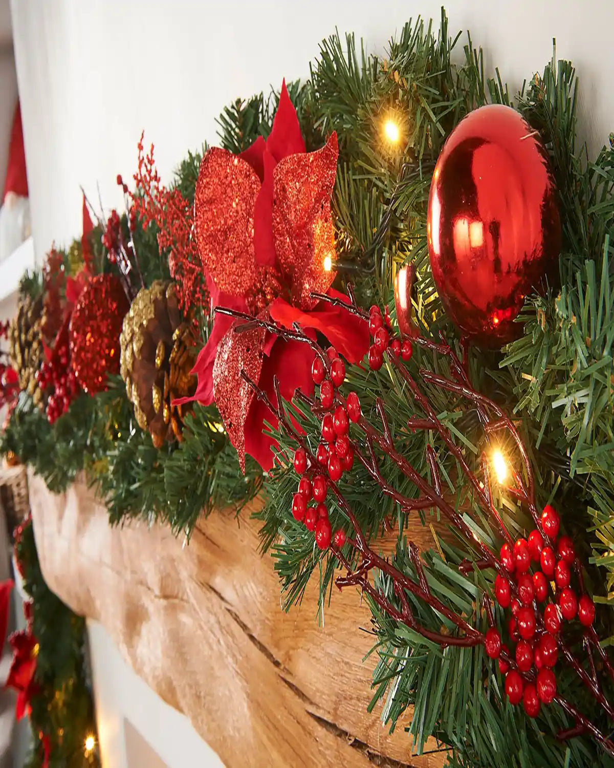 Close-up of a red and gold decorated garland on a wooden mantelpiece with a red bauble, berries, flowers and warm white lights