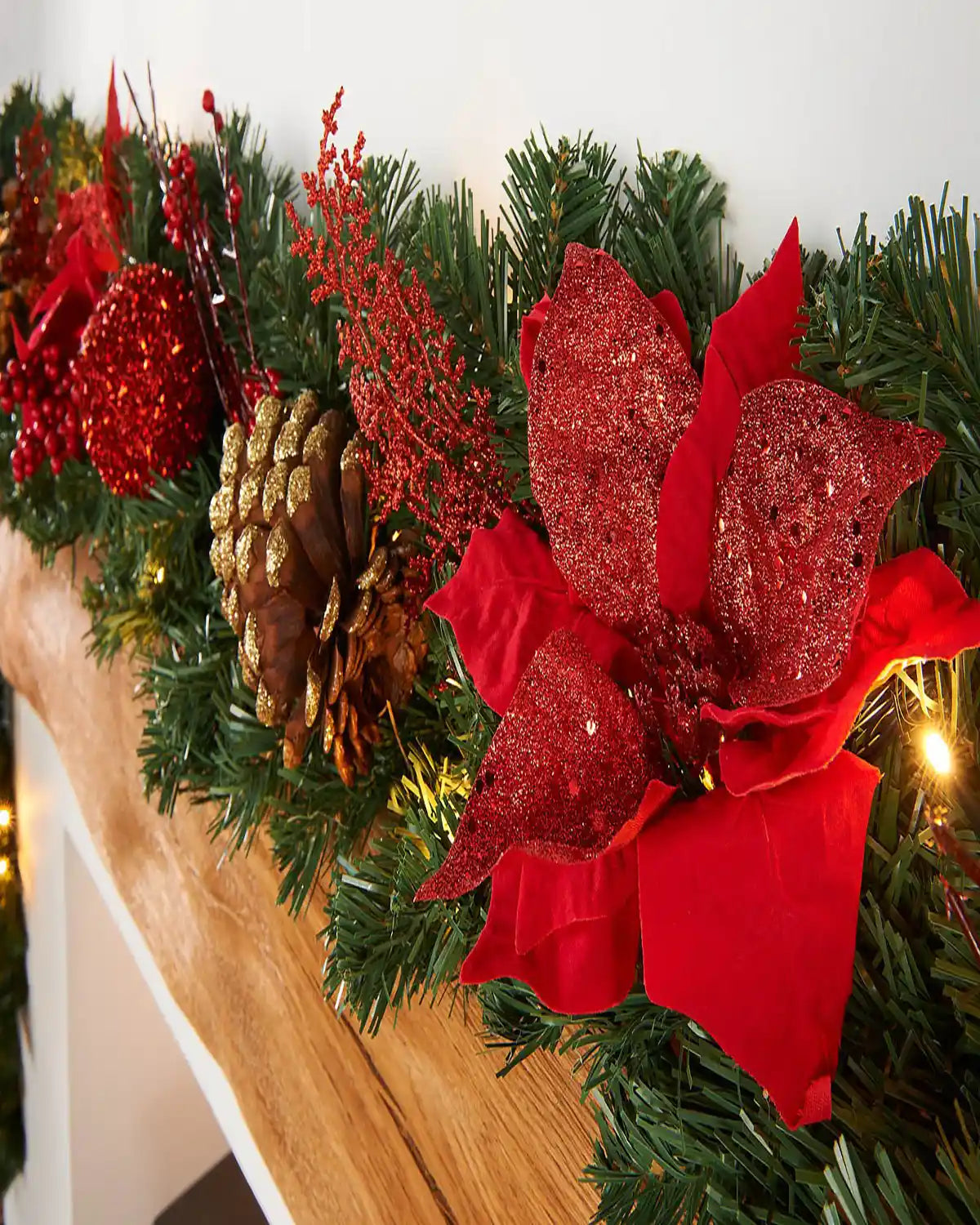 Close-up of a green Christmas garland with red flowers, red berries, gold-tipped pine cones and warm white lights