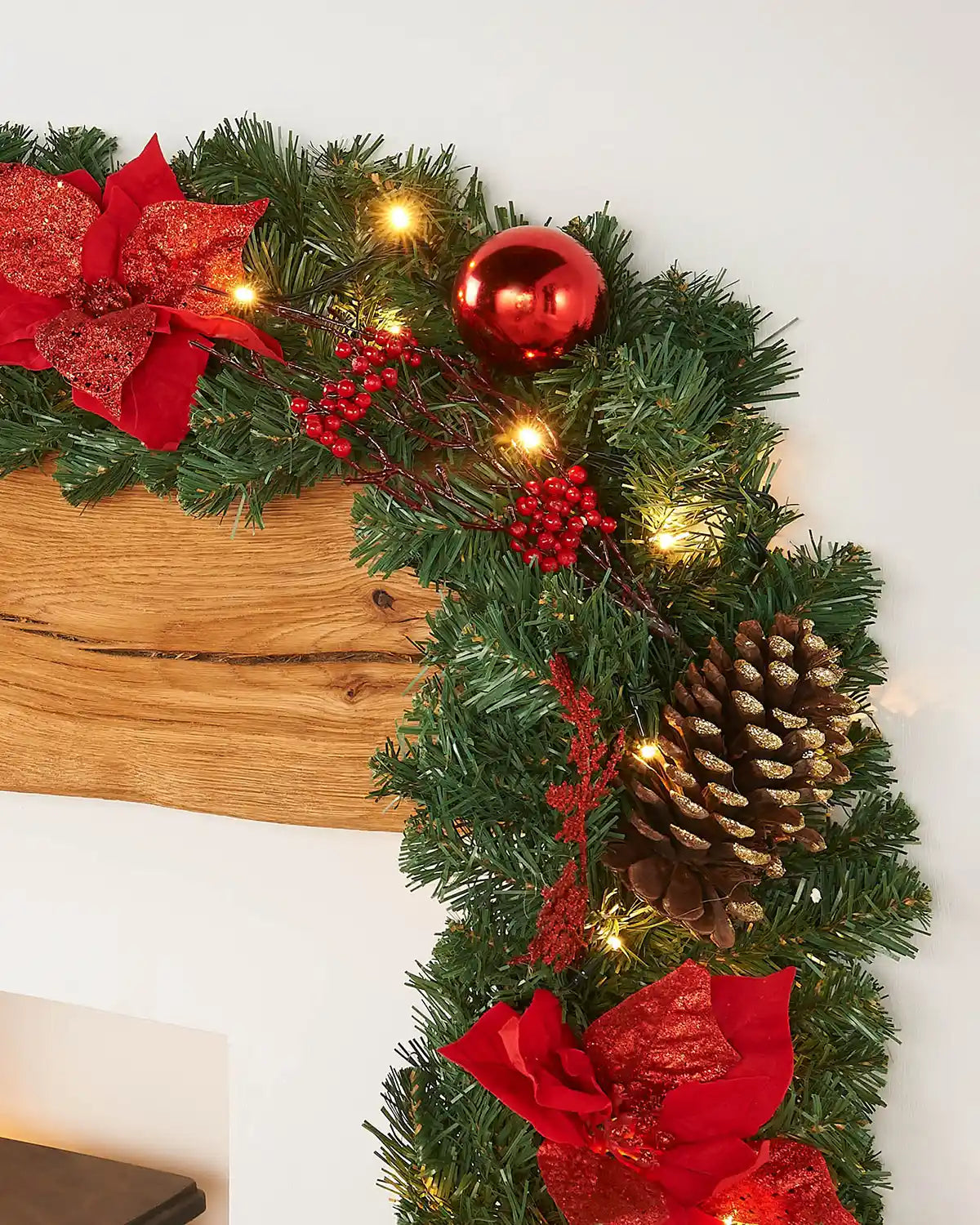 9ft pre-lit green garland on a wooden mantelpiece with red flowers, berries, a red bauble, pine cone and warm white lights