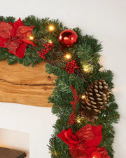 9ft pre-lit green garland on a wooden mantelpiece with red flowers, berries, a red bauble, pine cone and warm white lights