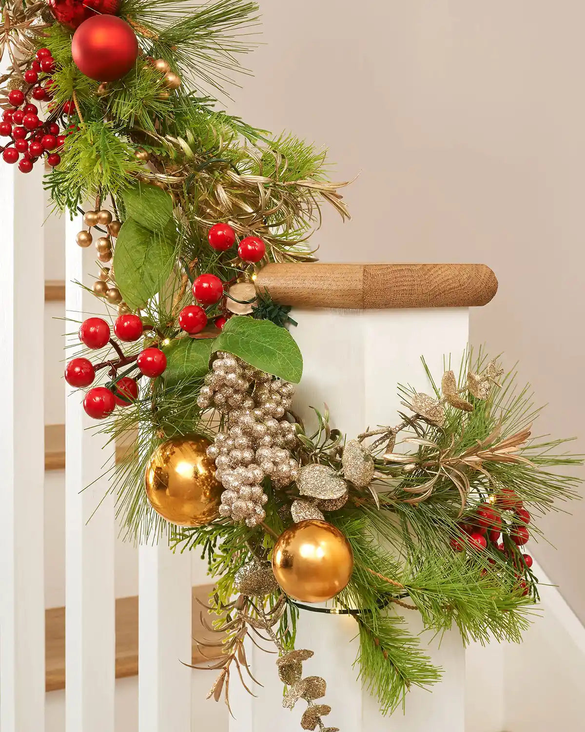 Detail of the 9ft pre-lit garland wrapped around a banister with red berries, gold baubles and mixed green foliage.
