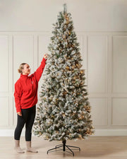 9 ft pre-lit slim snow flocked spruce Christmas tree with warm white lights and a person beside it for scale.