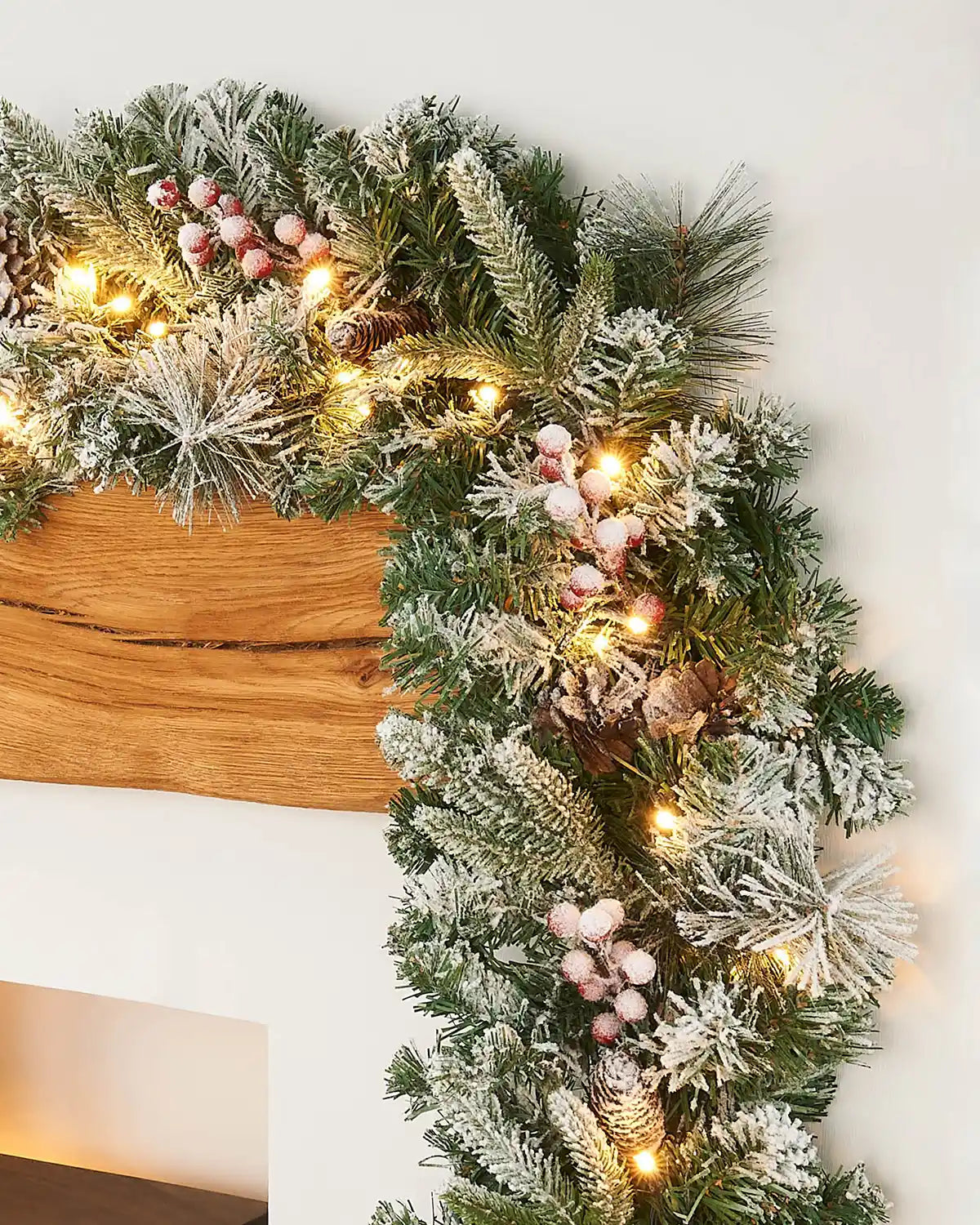Pre-lit snow flocked mixed pine garland displayed on a mantelpiece with warm white lights, pinecones and frosted berries