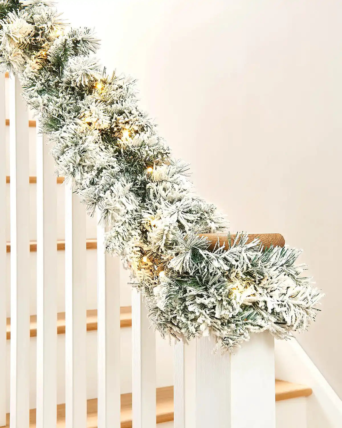 Snow-flocked spruce garland with warm white lights wrapped around a white stair banister