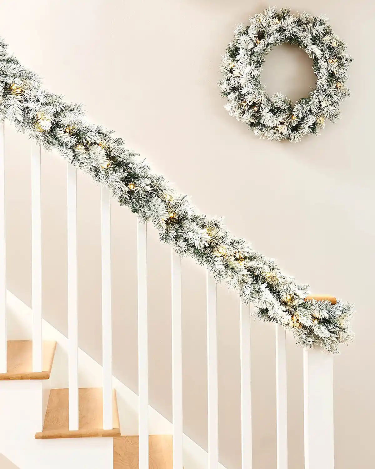 9ft pre-lit snow-flocked spruce garland draped along a white staircase with a matching wreath on the wall