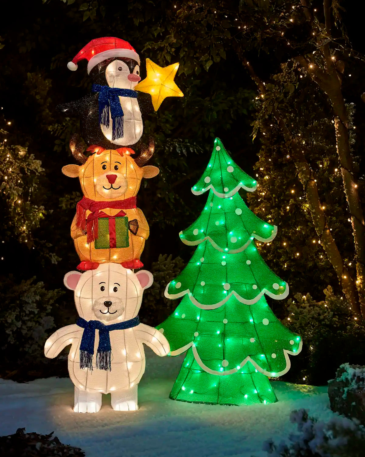 A  3D LED Polar bear with a reindeer standing on its shoulders with a penguin standing on the reindeers shoulders holding presents with the penguin putting a star on top of the Christmas tree against a dark garden setting on snowy grass and trees with lights in the background 