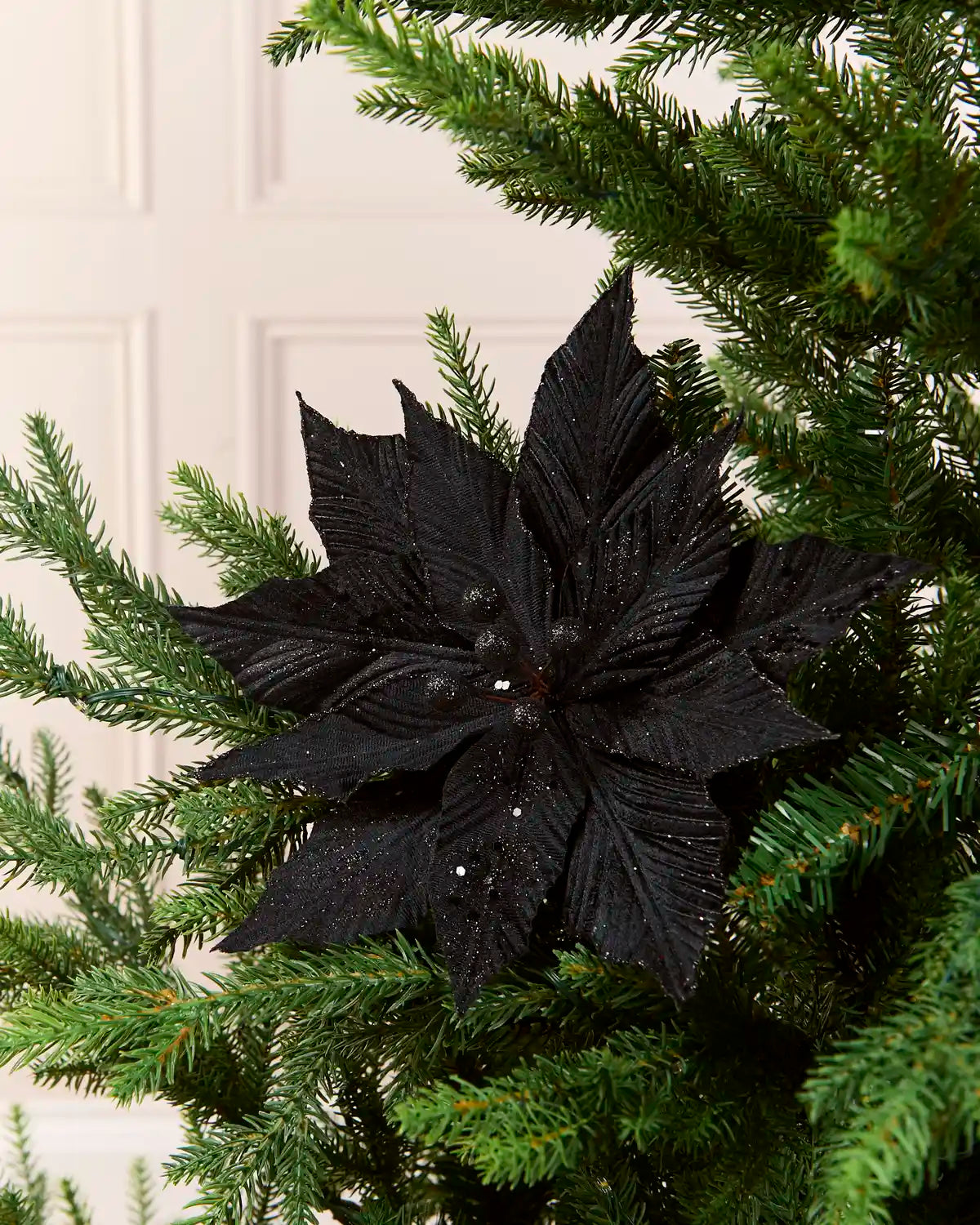 A black artificial poinsettia star ornament displayed on a green Christmas tree, with a soft-focus cream panneled door in the background.