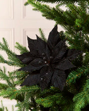 A black artificial poinsettia star ornament displayed on a green Christmas tree, with a soft-focus cream panneled door in the background.
