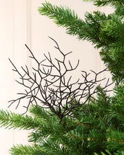 A black decorative coral style ornament with many thin twig-like branches, shown against a light cream background with green pine needles around it.