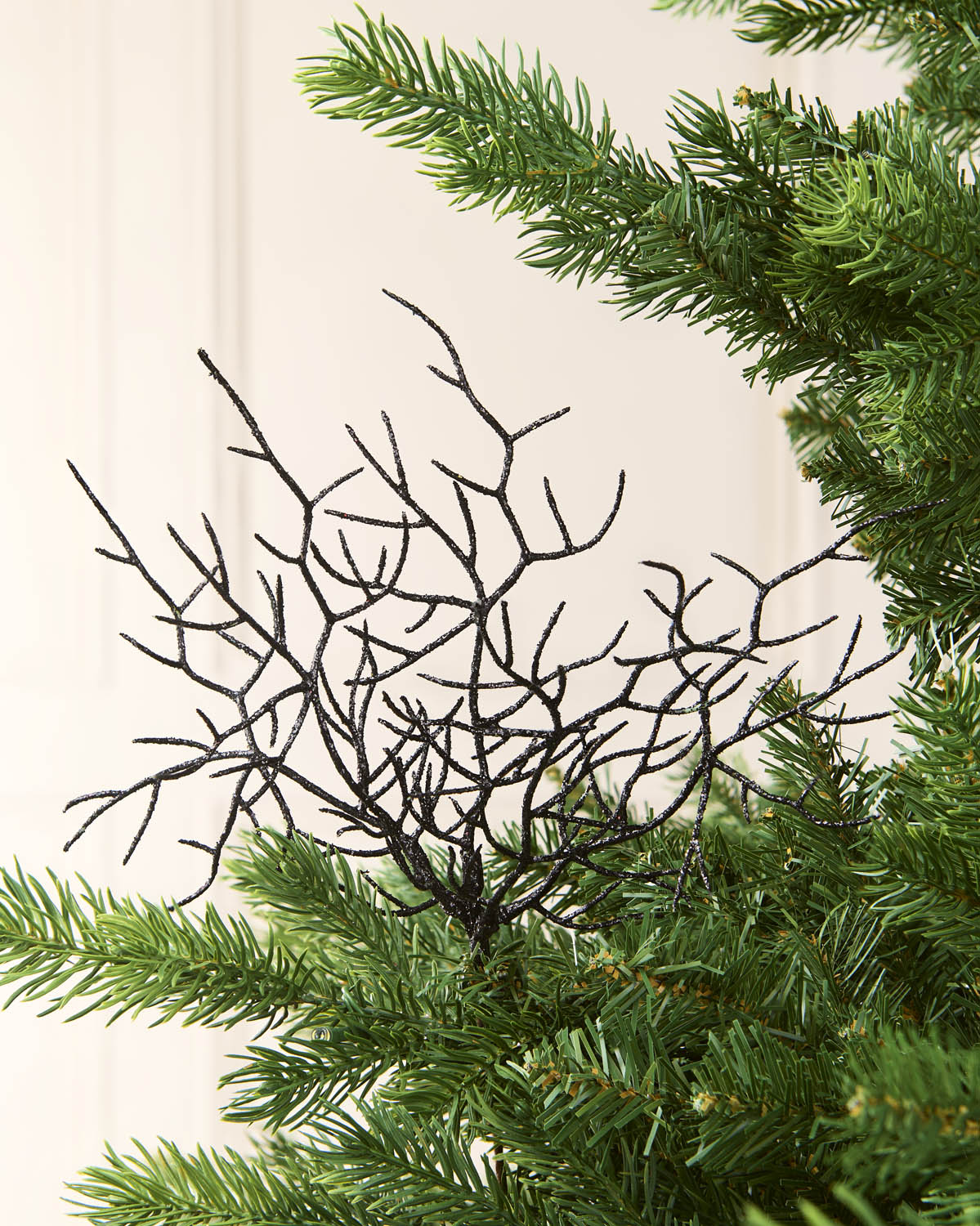 A black decorative coral style ornament with many thin twig-like branches, shown against a light cream background with green pine needles around it.