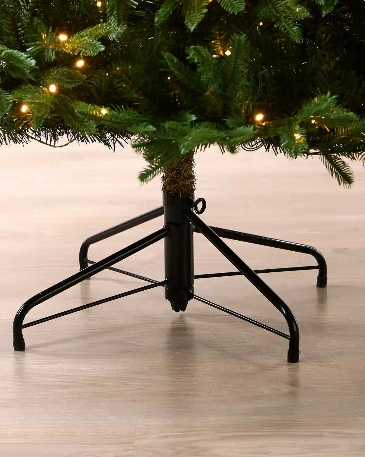 A black metal Christmas tree stand supporting a green artificial tree with warm white string lights, shown on a light wood floor.