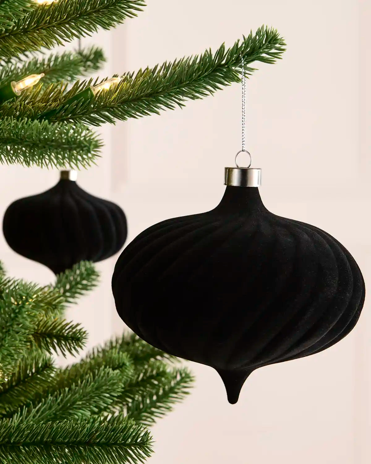 A black ribbed Christmas ornament hanging from a green pine tree branch with warm string lights in the background.
