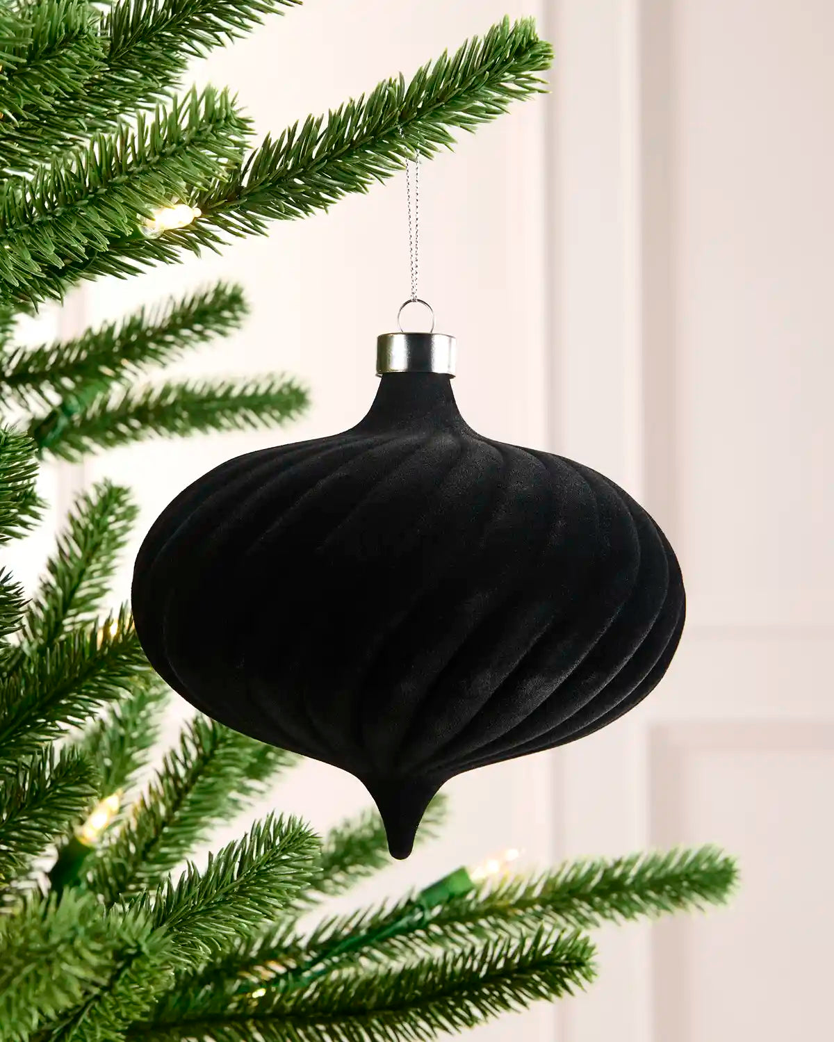 A black ribbed Christmas tree ornament hanging from a silver cap, suspended in front of green pine tree branches with warm white string lights.