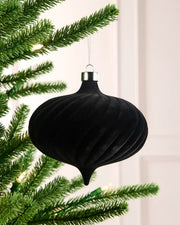 A black ribbed Christmas tree ornament hanging from a silver cap, suspended in front of green pine tree branches with warm white string lights.