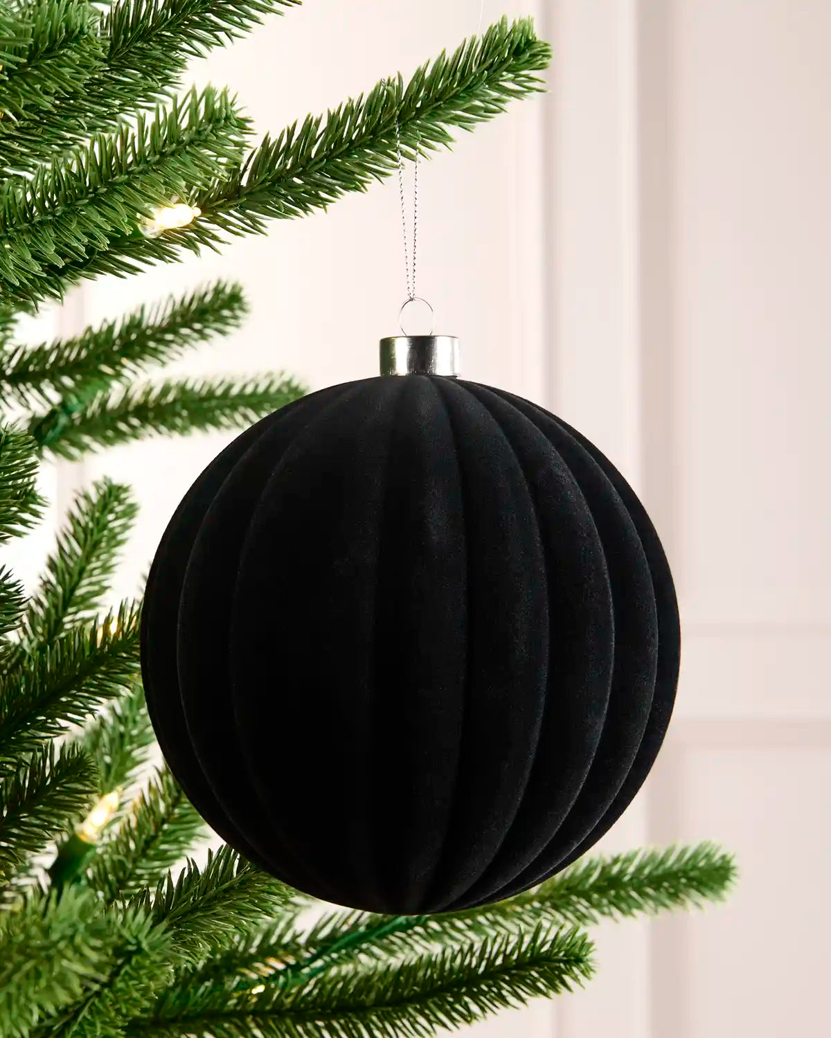 A black ribbed velvet Christmas ornament hanging from a green artificial tree branch with warm string lights in the background.