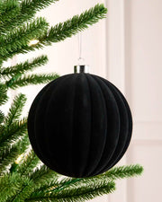 A black ribbed velvet Christmas ornament hanging from a green artificial tree branch with warm string lights in the background.