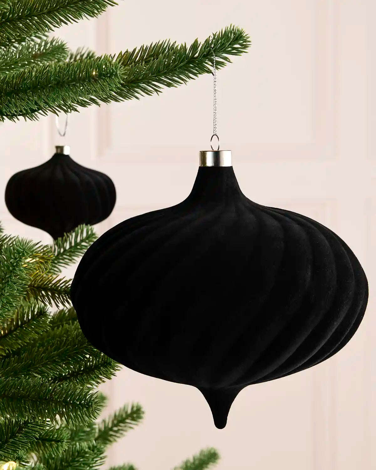 A black ribbed velvet Christmas ornament hanging from a green pine tree branch against a soft cream background, with a second ornament blurred in the background.