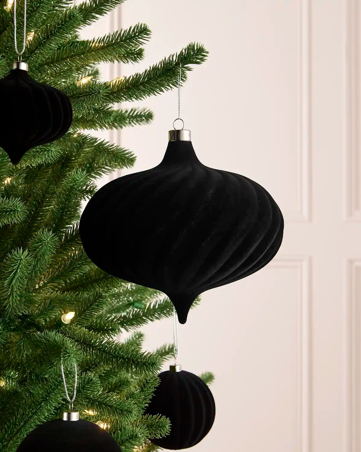 A black ribbed velvet Christmas tree ornament hanging from a green Christmas tree branch, with other matching black ornaments visible on the tree against a light cream panelled wall.