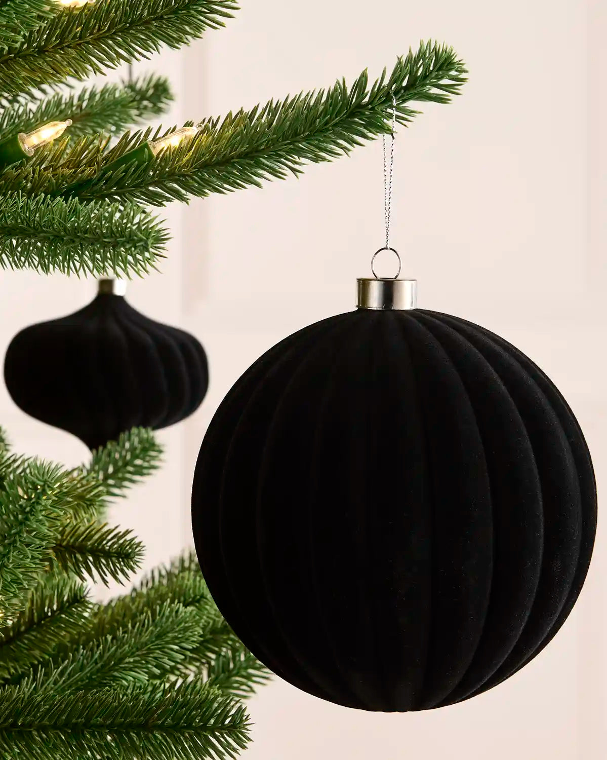 A black ribbed velvet Christmas tree ornament hanging from a green pine tree branch with warm string lights in the background.