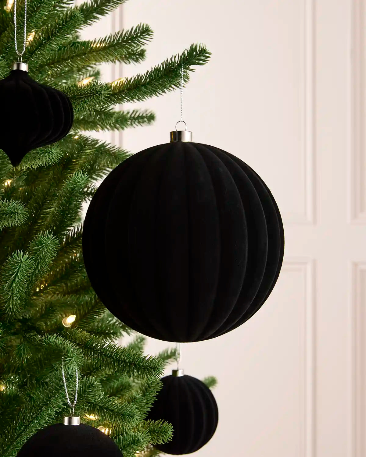 A black ribbed velvet Christmas tree ornament hanging on a green Christmas tree with warm white lights, against a light cream paneled wall.