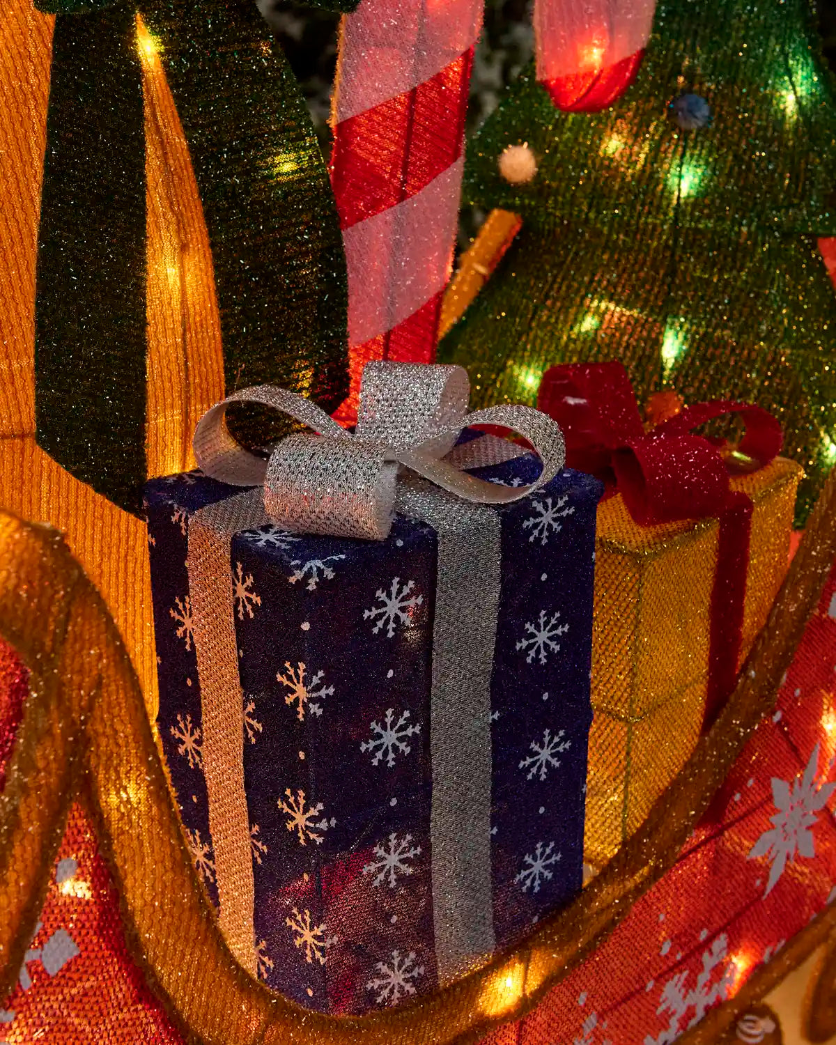 A close-up festive scene with a wrapped gift box in the foreground, decorated with a navy blue pattern of white snowflakes, a silver glitter ribbon, and a silver glitter bow. The gift box is surrounded by other holiday decorations, including a gold ornament, a red bow, a candy cane, and a Christmas tree with warm glowing lights.