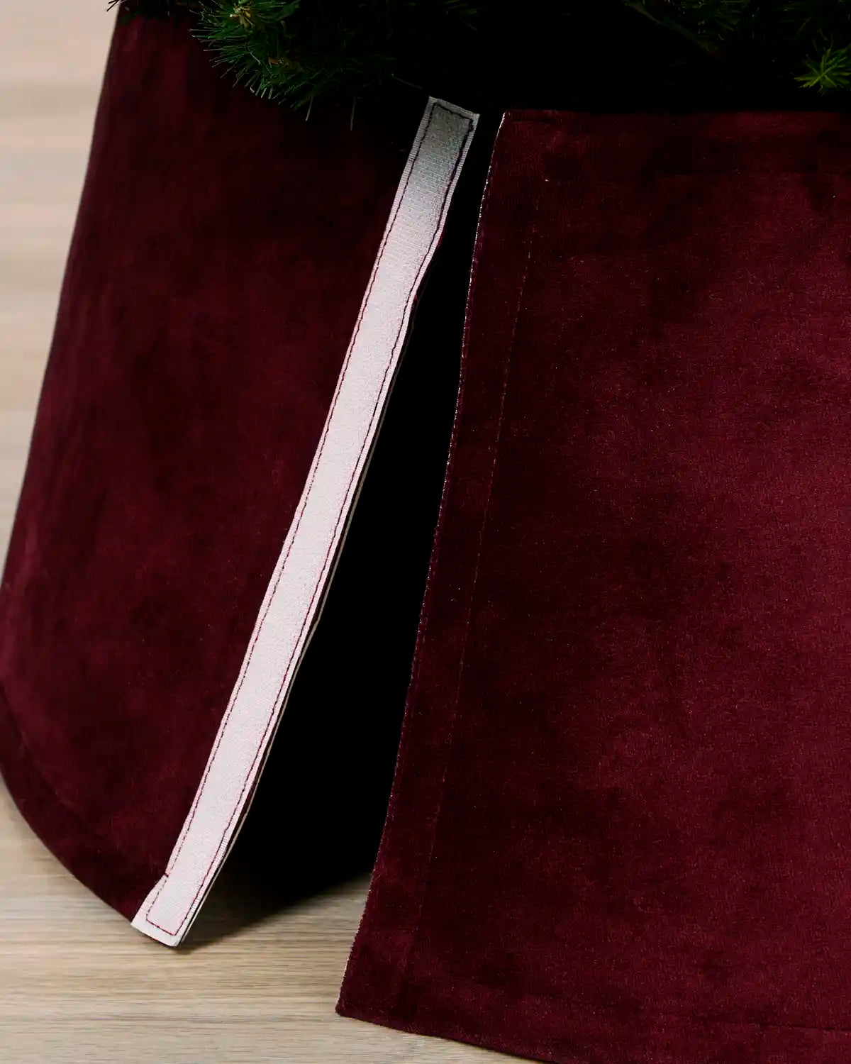 A close-up of a Burgundy coloured velvet Christmas tree skirt with a white trim, shown partially pulled open to reveal its shape and texture.
